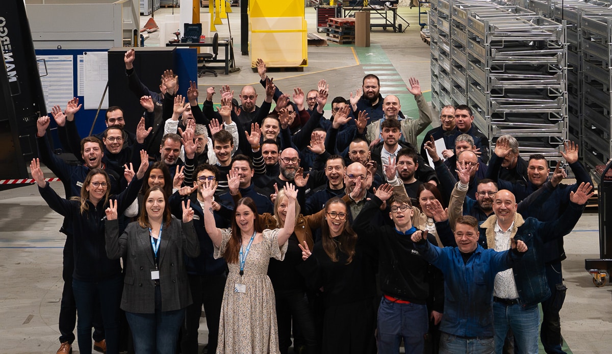 Groupe de collaborateurs et opérateurs de Manergo réunis, souriants et saluant joyeusement depuis l'atelier de fabrication."