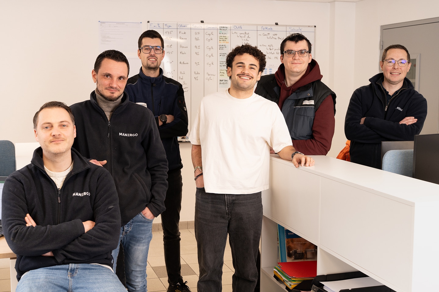 Six collaborateurs de l'équipe Manergo souriants, portant des vestes aux couleurs de l'entreprise dans leurs bureaux.