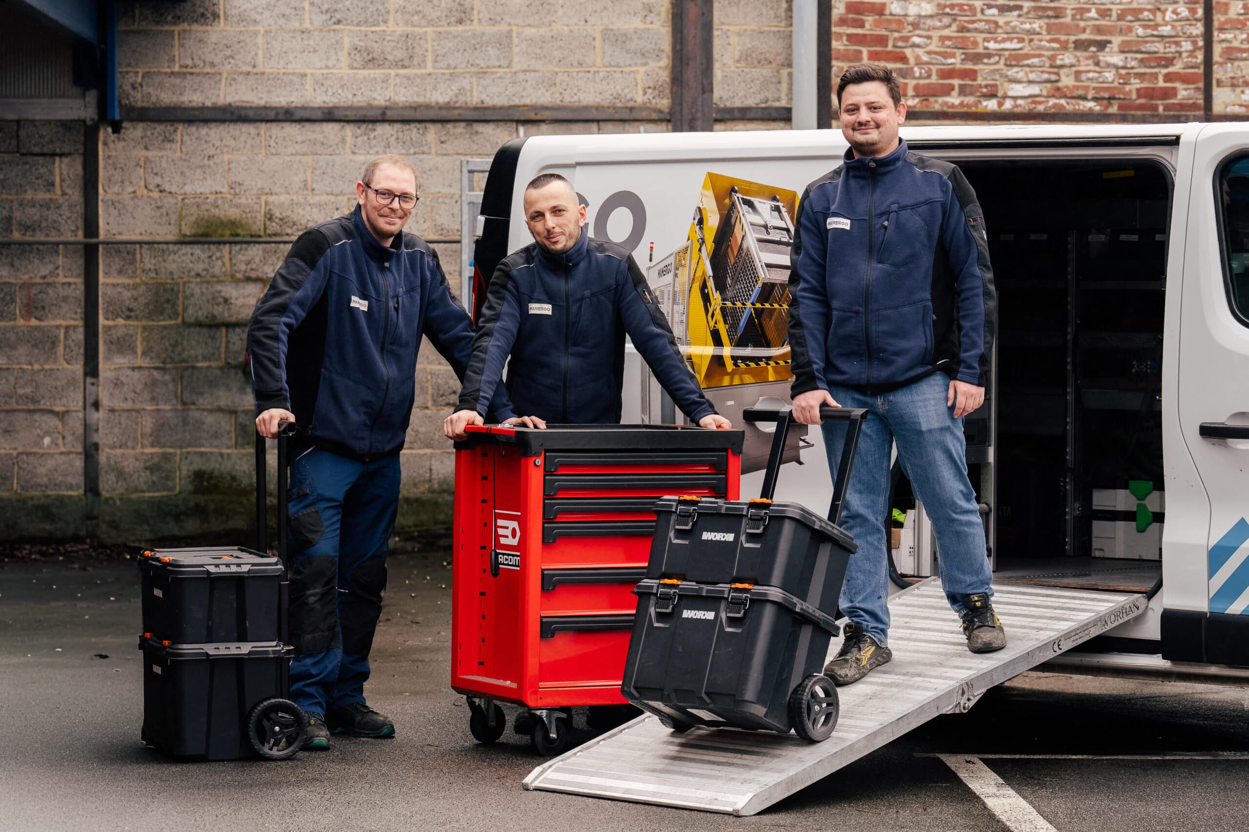 Trois techniciens du service maintenance de Manergo souriants à côté de leur véhicule d'intervention, équipés de servantes et de caisses à outils roulantes.
